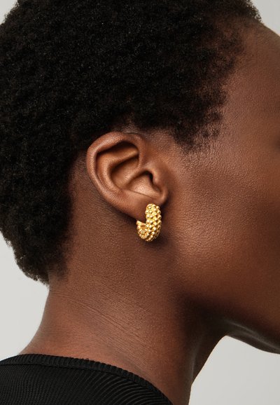 Közeli kép egy személy füléről, aki texturált arany karika fülbevalót visel, rövid, göndör hajjal és fekete felső látható.