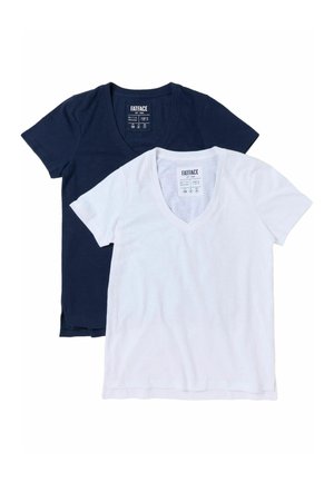 Ung kvinde med langt mørkt hår iført en mørkeblå T-shirt med V-udskæring og mørke shorts, stående mod en lys baggrund.