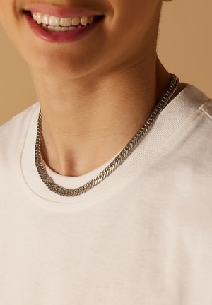 Zilveren ketting rustend op een lichtgekleurde t-shirt, met een dikke, geweven structuur en een gladde metalen afwerking.