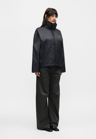 Γυναίκα όρθια, φοράει μαύρο μπουφάν με φερμουάρ και ψηλό γιακά και φαρδιά μαύρη παντελόνα, σε απλό ανοιχτό γκρι φόντο.