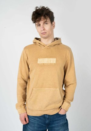 Sudadera beige con bolsillo tipo canguro, etiqueta bordada "GUESS LOS ÁNGELES", capucha con cordón y puños de punto. Textura de tela suave.