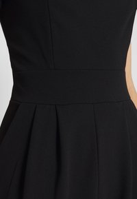 Robe noire avec un corsage ajusté, une large ceinture à la taille et une jupe plissée. Texture lisse avec des coutures discrètes le long des coutures.