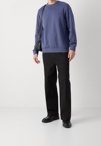 Lila sweatshirt, rund hals, ribbade muddar, kombinerad med svarta byxor och skor. Enkel design, tygtextur synlig, liten logotypdetalj.