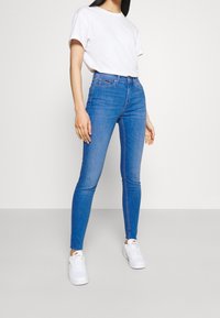 Jeans de mezclilla azul de tiro alto con un corte ajustado, combinados con una camiseta blanca lisa y zapatillas blancas. Textura suave, sin patrones notables.