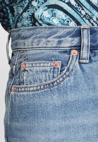 Jeans in denim azzurro chiaro con bordi sfilacciati, bottone e rivetti color rame e un classico design a cinque tasche. Cuciture intricate sulla cintura.