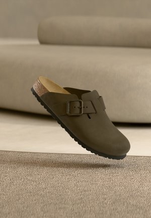 Brązowy chodak wsuwany z dużą klamrą i korkową wkładką unoszący się nad beżowym wykładzinowym podłożem w minimalistycznym pomieszczeniu.