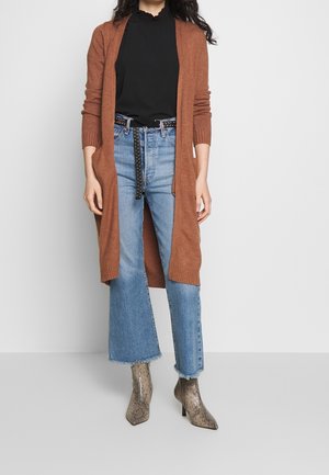 Femme portant un long cardigan marron, un haut noir, un jean court bleu avec ourlet effiloché, une ceinture cloutée et des bottines imprimé serpent.