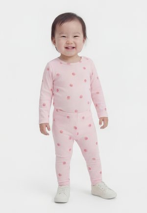 Malé dievčatko sa usmieva, má na sebe ružový dlhý rukáv a legíny so vzorom drobných červených jabĺk a biele tenisky na bielom pozadí.