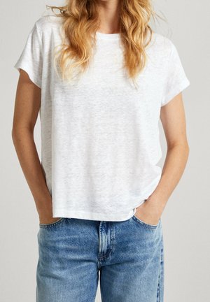 Blonde person wearing a plain white short-sleeve t-shirt tucked slightly into light blue denim jeans with hands in pockets.