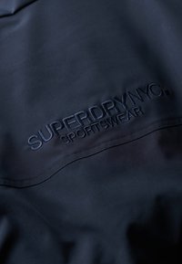 Granatowa marynarka sportowa z haftowanym logo "SUPERDRY NYC SPORTSWEAR". Gładka faktura z warstwowym wzorem na tkaninie.