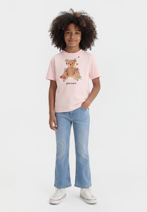 Enfant aux cheveux bouclés portant un t-shirt rose avec un motif d'ours en peluche, un jean bleu clair et des baskets blanches, debout, une main dans la poche.