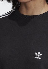 Čierna mikina so strihaným okrajom výstrihu, so bielym logom Adidas a tromi bielymi pruhmi na pleci. Mäkká textúra látky.