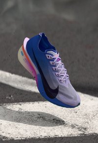 Μπλε και μοβ παπούτσι τρεξίματος Nike με μαύρο σήμα αιωρείται πάνω από άσφαλτο με λευκές βαμμένες σημάνσεις.