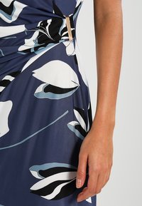 Robe bleu marine à motif floral blanc et noir, avec un design torsadé et un accent en métal doré à la taille. Texture de tissu lisse.