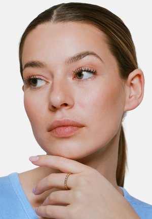 Frau mit leichtem Make-up und zurückgebundenen Haaren, trägt ein hellblaues Oberteil, stützt das Kinn auf die Hand mit einem goldenen Ring am Mittelfinger.