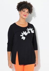 Top lungo nero a maniche lunghe con applicazioni di fiori bianchi e spacchi laterali. Tessuto morbido, scollatura rotonda e vestibilità comoda. Abbinato a pantaloni arancioni.