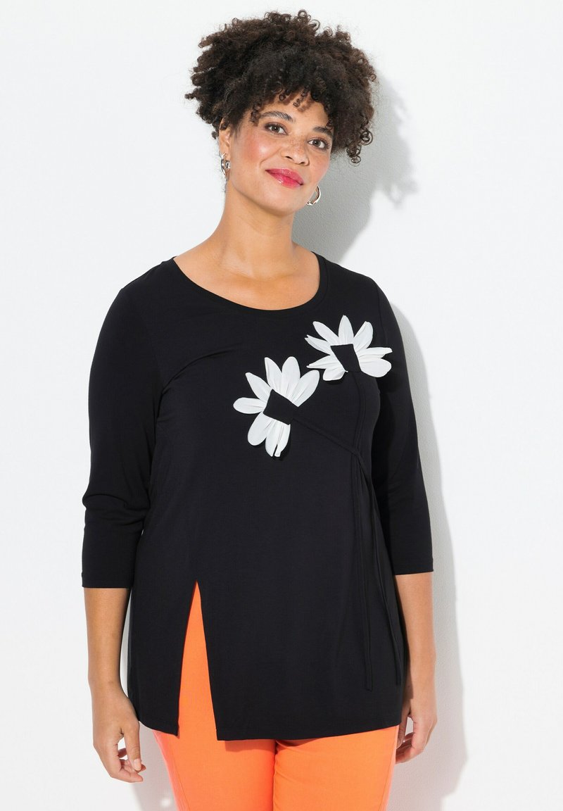 Top lungo nero a maniche lunghe con applicazioni di fiori bianchi e spacchi laterali. Tessuto morbido, scollatura rotonda e vestibilità comoda. Abbinato a pantaloni arancioni.