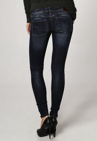 Jeans ajustados de color azul oscuro con una textura deslavada, que cuentan con dos bolsillos en la parte posterior y un parche de cuero en la cinturilla, combinados con tacones altos negros.
