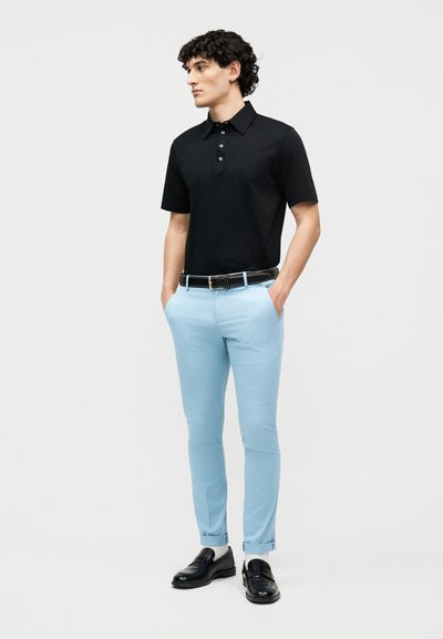 Νέος άνδρας όρθιος με τα χέρια στις τσέπες, φοράει μαύρο πόλο με κοντά μανίκια, ανοιχτό μπλε παντελόνι στενής γραμμής, μαύρη ζώνη, λευκές κάλτσες και μαύρα μοκασίνια.