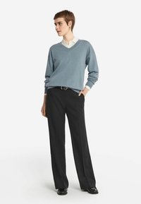 Jasnoniebieski sweter z dekoltem w serek, wykonany z miękkiego materiału, w połączeniu z czarnymi szerokimi spodniami i czarnymi butami. Pod spodem widoczna biała koszula z kołnierzykiem.