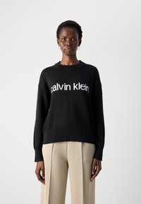 Schwarzer Strickpullover mit dem weißen "calvin klein" Logo, runder Ausschnitt, fallende Schultern und geripptem Saum. Kombiniert mit beigen Hosen.
