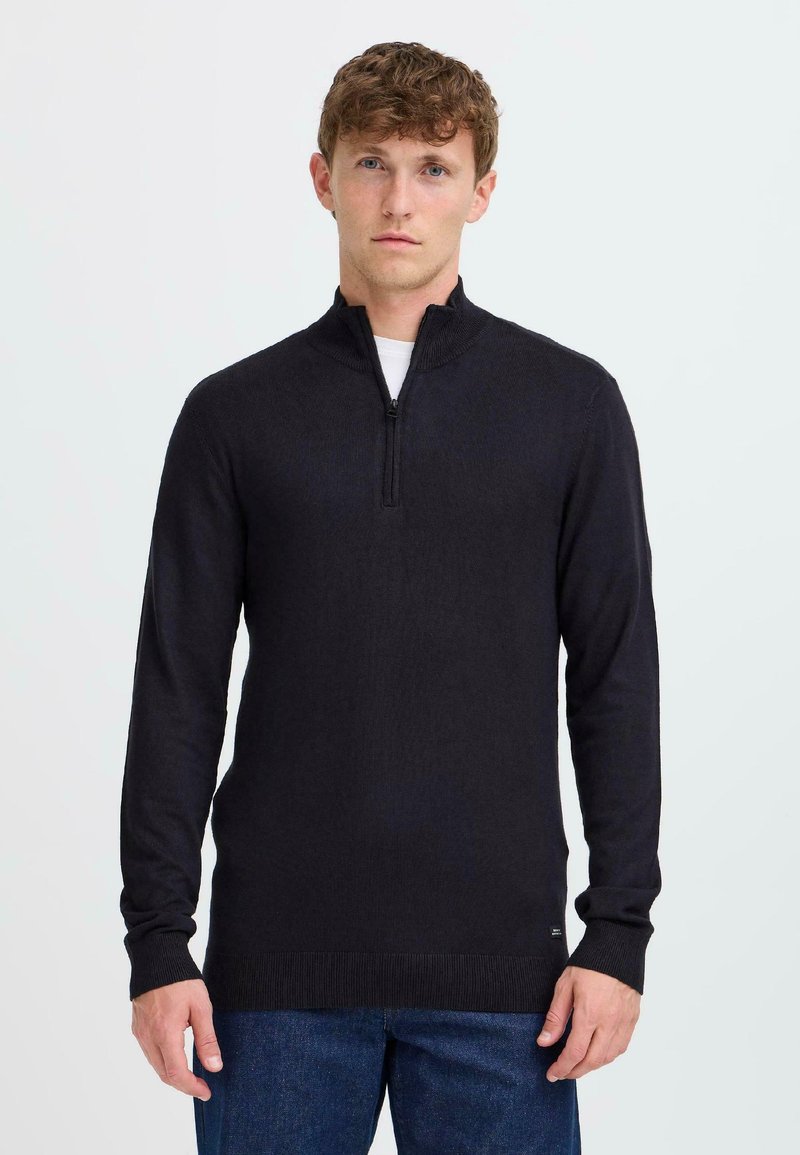 Pull à col zippé noir avec des manches longues et des poignets côtelés, fabriqué en matériau tricoté doux. Présente une texture lisse et une coupe ajustée.