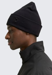 Zwarte gebreide beanie met een geribbelde textuur, gekenmerkt door een subtiele logo aan de voorkant. Draag op het hoofd van een persoon in een donkere jas.