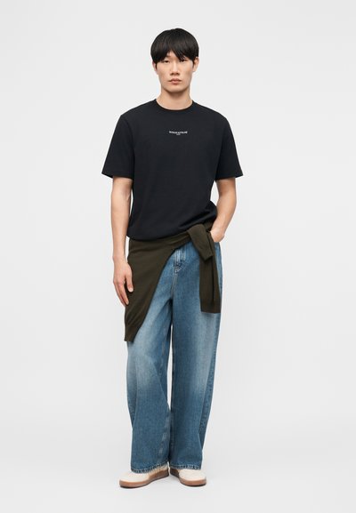 Νεαρός άντρας που φοράει μαύρο T-shirt Maison Kitsuné, φαρδιά μπλε τζιν, λευκά παπούτσια και ένα χακί πουλόβερ δεμένο γύρω από τη μέση του.