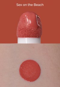 Colore labbra liquido in una tonalità corallo lucida su un applicatore a spugna e una macchia circolare sulla pelle. La texture appare liscia e cremosa.