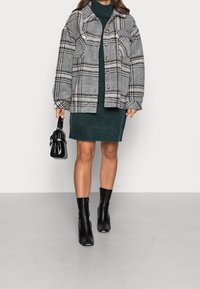 Giacca a quadri grigia con tasche laterali, cinque bottoni in legno, abbinata a un vestito maglione verde scuro e stivali neri alla caviglia. Porta una piccola borsa nera.