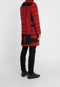 Czerwony płaszcz z mieszanki wełny z czarnymi paskami i dużym logo. Posiada dopasowany krój, długie rękawy i wysoki kołnierz. Nosi się go na czarne spodnie i buty.