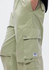 Ανοιχτό πράσινο παντελόνι cargo με υφή υφάσματος. Διαθέτει μεγάλες πλαϊνές τσέπες, κουμπιά με στερεώσεις και ετικέτα με λογότυπο στην μπροστινή τσέπη.