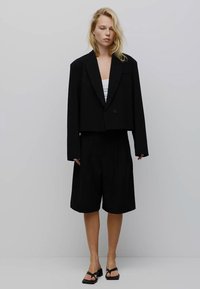 Blazer cropped nero con un bottone, abbinato a pantaloni corti pieghettati neri. La modella indossa una maglietta bianca e sandali neri. Sfondo neutro.