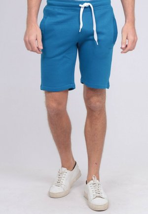 Homme portant un short bleu à cordon et des baskets blanches, debout devant un fond clair uni.