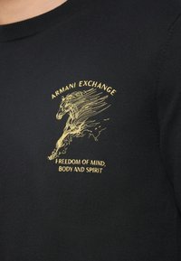 Μαύρο πουκάμισο με χρυσή κεντημένη κεφαλή αλόγου, "ARMANI EXCHANGE" από πάνω και "ΕΛΕΥΘΕΡΙΑ ΝΟΥ, ΣΩΜΑΤΟΣ ΚΑΙ ΠΝΕΥΜΑΤ�ΟΣ" κάτω από το σχέδιο.