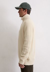 Beige gebreide trui met een hoge kraag, voorzien van geribbelde manchetten en zoom. Gedragen met een groene pet, getoond tegen een effen achtergrond.