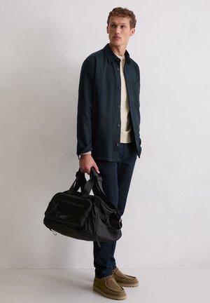 Mężczyzna w ciemnej kurtce i spodniach trzyma czarną torbę sportową, nosi jasny sweter i brązowe zamszowe buty, stoi przy białej ścianie.