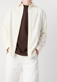 Camicia button-up in corda crema sopra una maglietta marrone, abbinata a pantaloni bianchi. Presenta un colletto e un logo discreto sul petto.