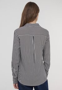 Camicia a maniche lunghe a righe bianche e nere con colletto, caratterizzata da una piega sul retro e un orlo arrotondato, realizzata in tessuto liscio.