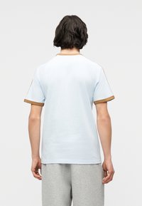 Άτομο με κοντά σκουρόχρωμα μαλλιά που φοράει ένα ανοιχτό μπλε πουκάμισο με κοντά μανίκια και καφέ περιθώριο, καθώς και γκρι παντελόνι, γυρισμένο με την πλάτη προς το απλό φόντο.