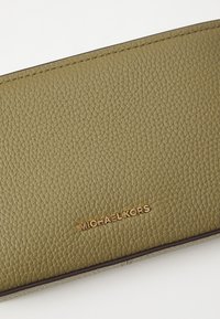 Olive vihreä nahkalompakko, jossa on karkeapintainen rakenne, ja jossa on kultaiset "MICHAEL KORS" -brändäykset sekä tummanruskea reunus. Yksinkertainen, suorakulmainen muotoilu.