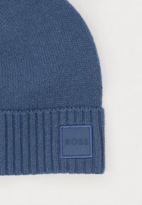 Căciulă tricotată albastră cu mansetă ondulată, având un eticheta texturată de formă pătrată cu "BOSS" embosat pe ea. Material moale, texturat.