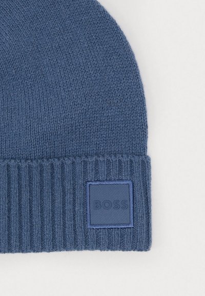 Bonnet en tricot bleu avec un revers côtelé, arborant une étiquette carrée texturée avec le mot "BOSS" embossé dessus. Tissu doux et texturé.