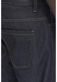 Närbild på mörkblå denimjeans med synliga gula sömmar och detaljer på bakfickan vid en persons höft.