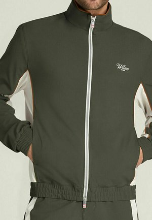 Uomo che indossa una giacca con cerniera verde scuro e bianca, con polsini e orlo elasticizzati, mani nelle tasche, logo del marchio Wilson sul petto.