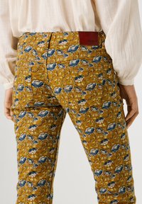 Παντελόνια με λουλουδένιο μοτίβο σε μουσταρδί κίτρινο, με μπλε τριαντάφυλλα, πράσινα φύλλα και κόκκινο δερμάτινο ετικέτα επωνυμίας στη πίσω ζώνη μέσης.