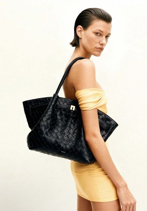 Femme aux cheveux courts plaqués, portant une robe jaune épaules dénudées, portant un grand sac cabas en cuir noir tressé sur l'épaule.