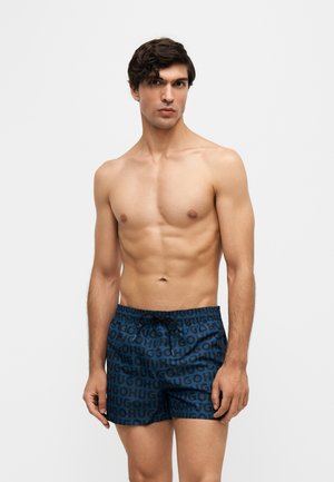Jeune homme aux cheveux courts et foncés portant un short de bain bleu avec un texte imprimé noir "HUGO" sur un fond clair uni.