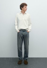 Hvid langæret polo med tre-knaps placket, parret med afslappede, falmede denim jeans og brune ruskindssko, stående på grå gulv.