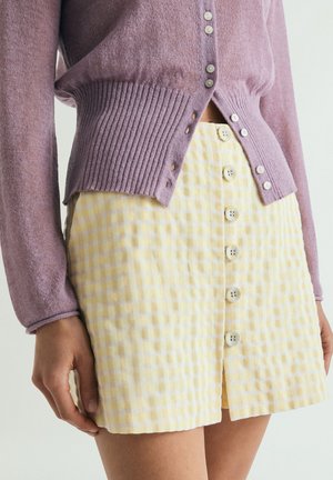 Persona que lleva un cárdigan lila con botones sobre una falda de botones de talle alto con cuadros amarillos y blancos.